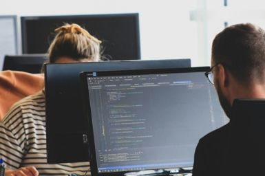 Two people working on monitors facing each other, with code on the screen, photo by Sigmund on Unsplash