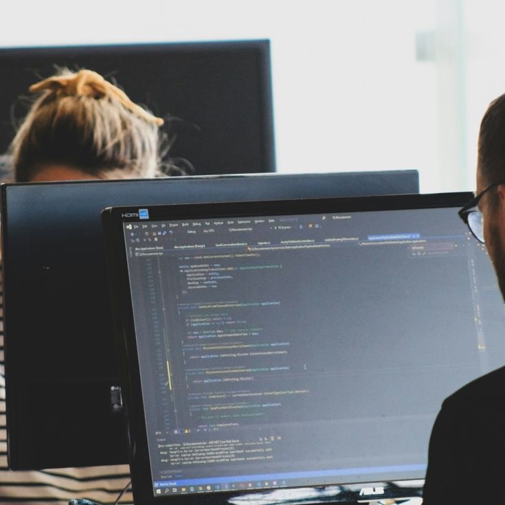 Two people working on monitors facing each other, with code on the screen, photo by Sigmund on Unsplash